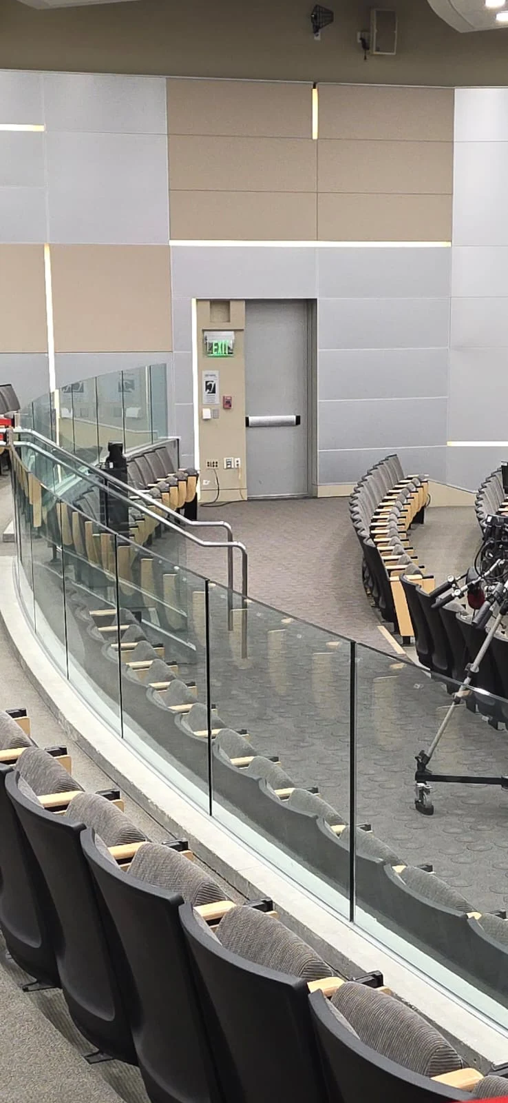 A close view of the clear glass barrier and stainless steel handrail along the auditorium’s stepped seating.