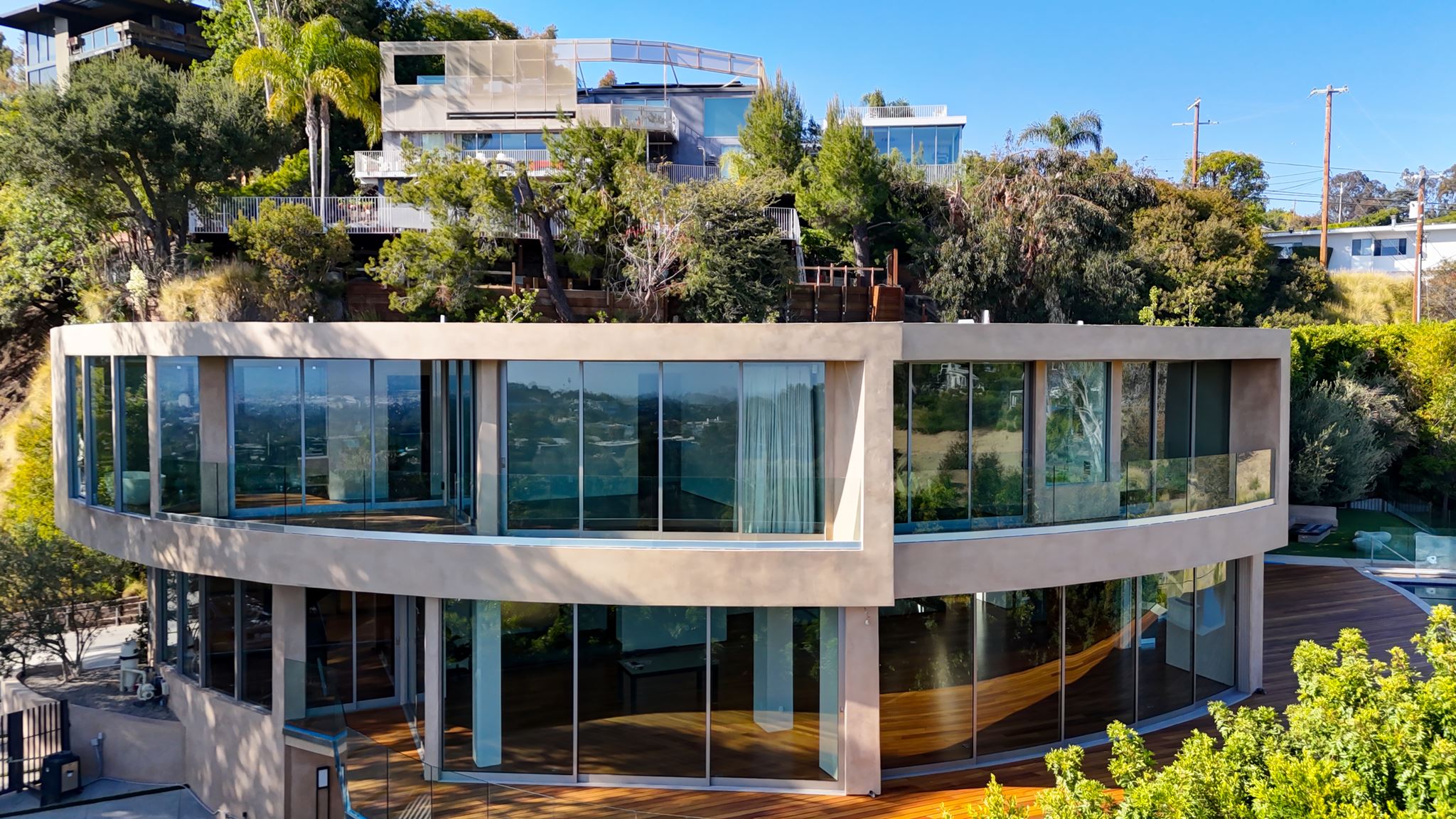 Curved, floor-to-ceiling glazing wrapping a two-storey hillside residence in Brentwood, seen from above with the Los Angeles basin behind.